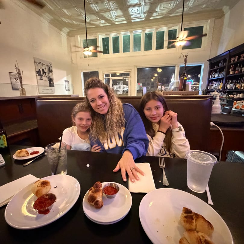 A woman and two girls pose at a restaurant table with breadsticks and dipping sauce.