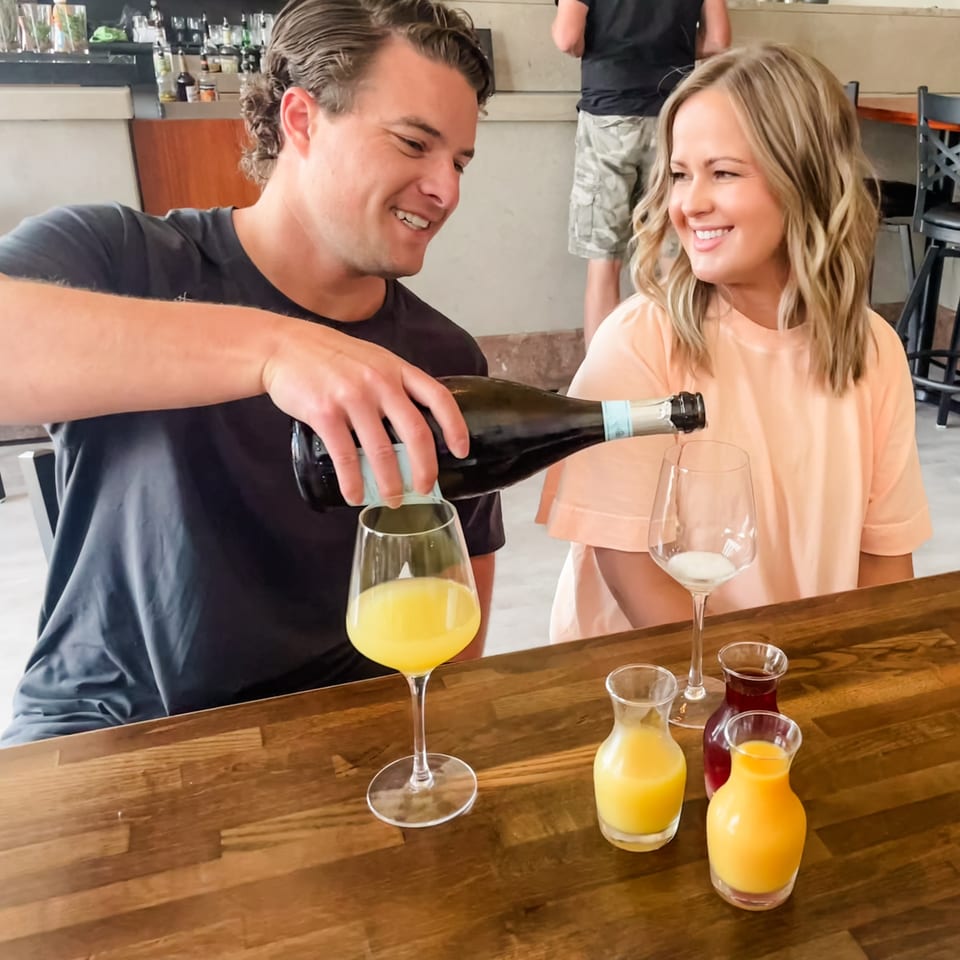 Man pours sparkling wine for mimosas as a woman smiles at a restaurant table.
