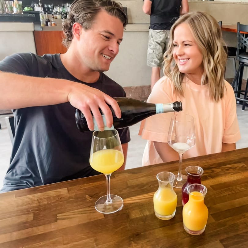 Man pours sparkling wine for mimosas as a woman smiles at a restaurant table.