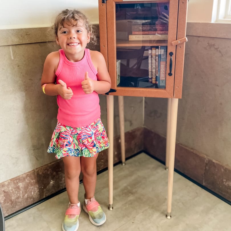 Smiling young girl in a pink top and floral skirt gives two thumbs up next to a small bookshelf.