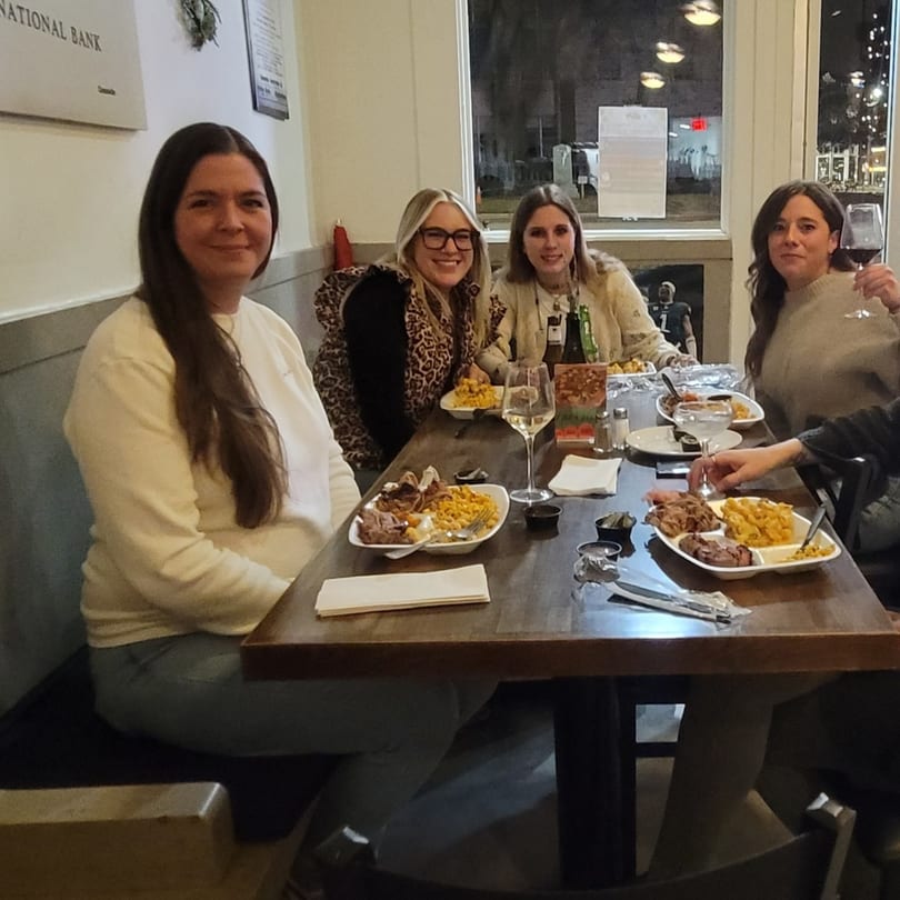 Four women enjoying dinner and drinks at a restaurant table with plates of food.