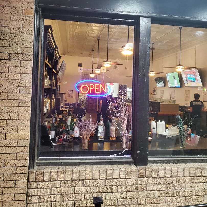 Exterior view of a restaurant window showing an "OPEN" neon sign and a well-stocked bar inside.
