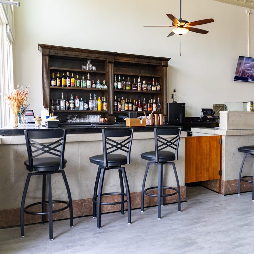 Empty bar area with black stools, a dark wood liquor shelf, and a ceiling fan.