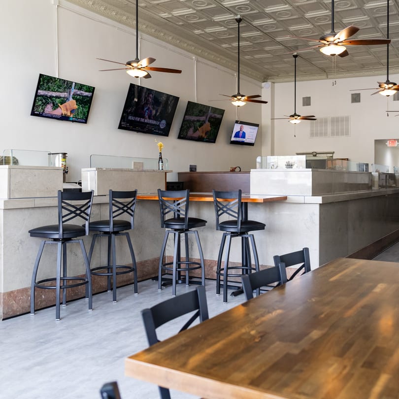 Modern restaurant interior featuring a bar with stools, dining tables, wall-mounted TVs, and ceiling fans.