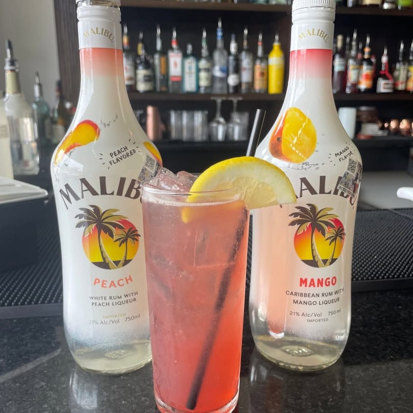 Pink cocktail with lemon slice flanked by Malibu Peach and Mango rum bottles on a bar.