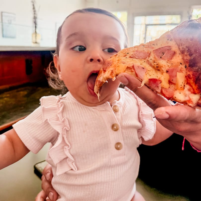 A baby with wide eyes eagerly takes a bite of a pizza slice held by an adult hand.
