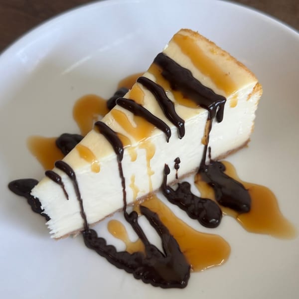 A slice of creamy cheesecake with chocolate and caramel drizzles on a white plate.