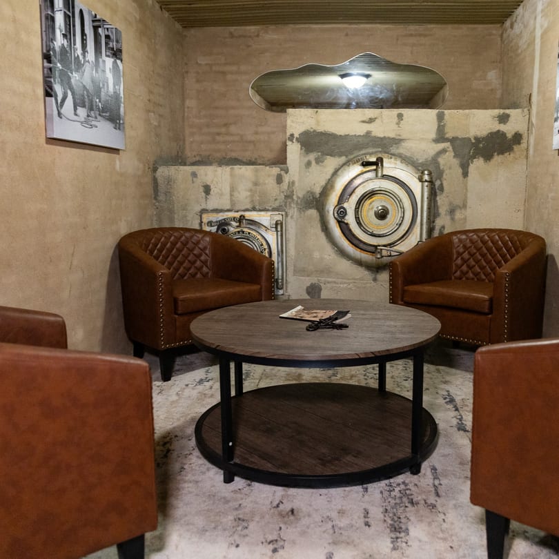 Cozy lounge with brown leather chairs, a wooden table, and bank vault decor on textured walls.