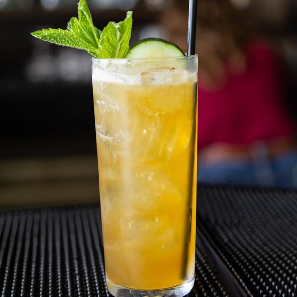 A vibrant yellow cocktail with ice, garnished with fresh mint and a cucumber slice, on a bar mat.