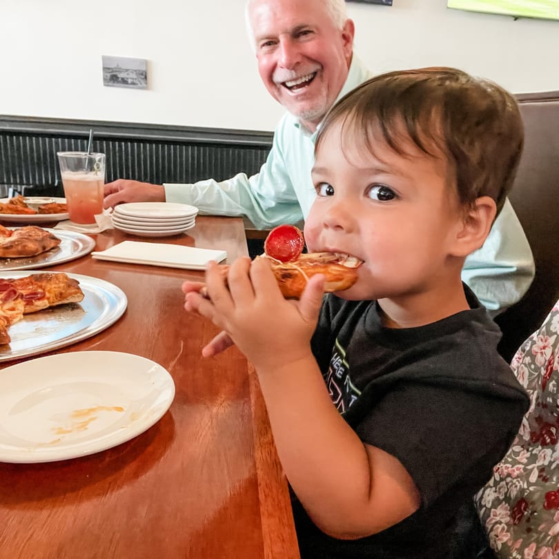 Smiling child eats a folded pizza slice at a restaurant table, with a happy man behind.