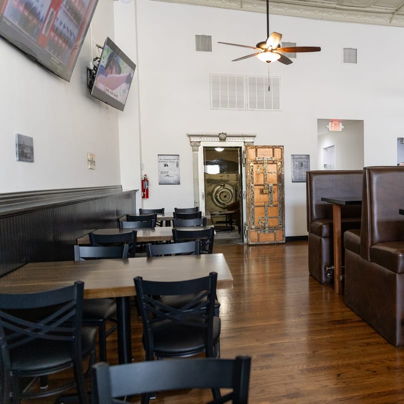 Restaurant interior with wooden tables, black chairs, brown booths, and an open bank vault door.