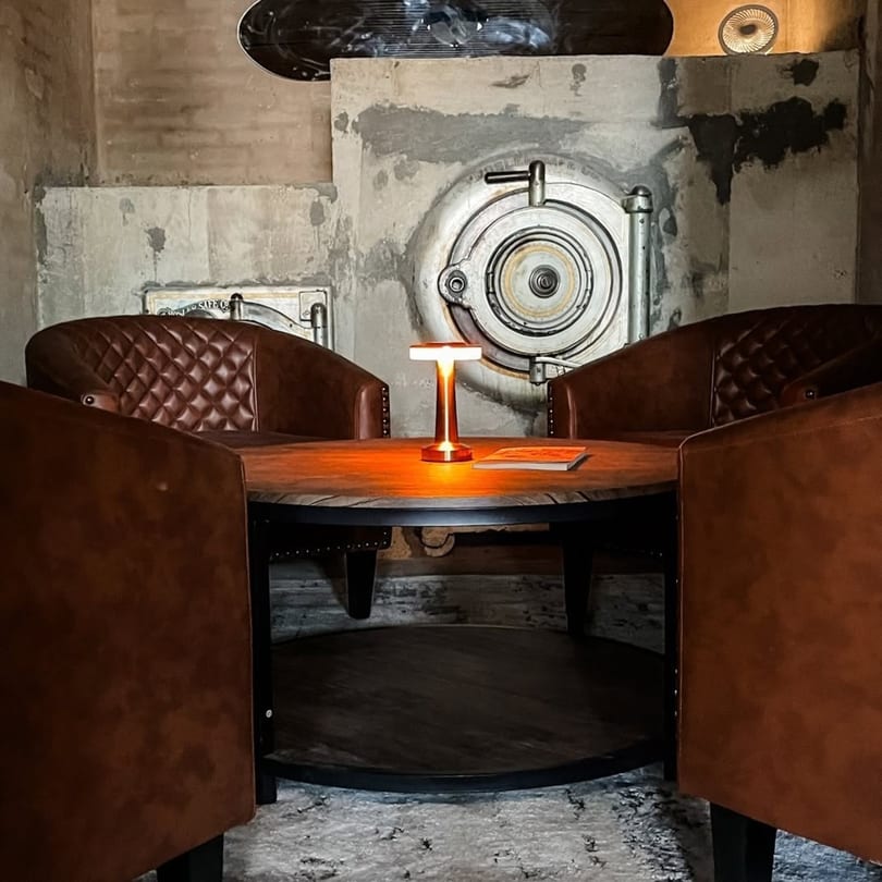Industrial-chic lounge area featuring brown leather armchairs, a round table, and a large vault door.