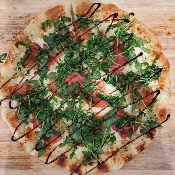 Sliced pizza with white cheese, prosciutto, fresh arugula, and balsamic glaze, on a light wooden board.