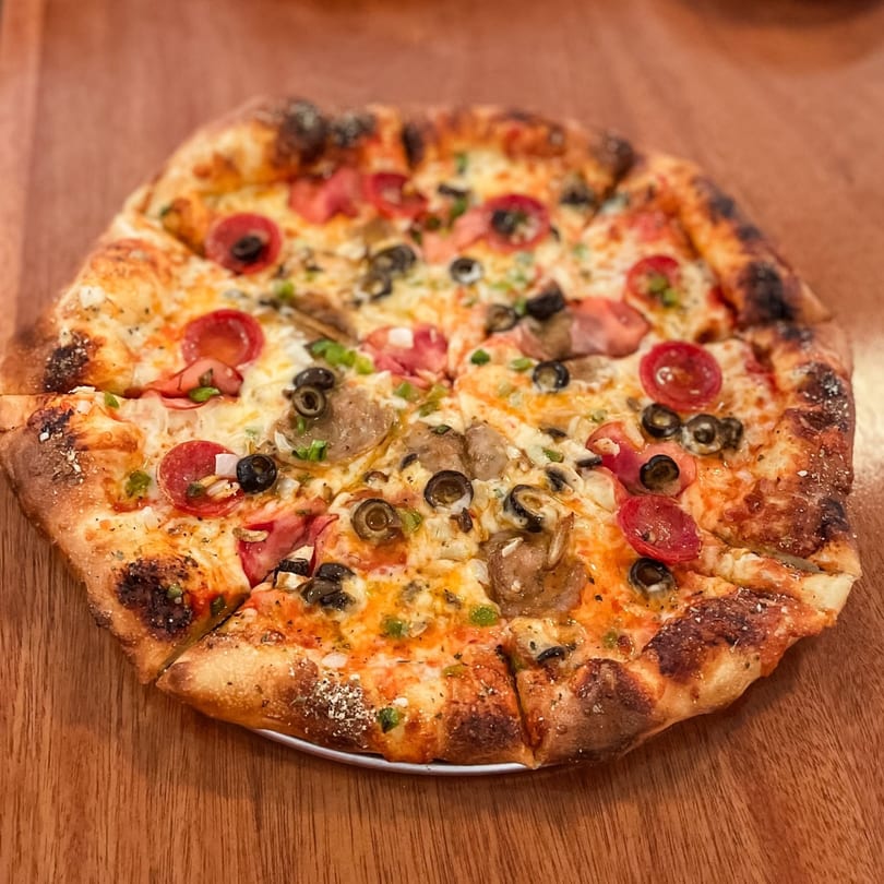 Whole pizza with pepperoni, olives, mushrooms, and peppers on a wooden table.