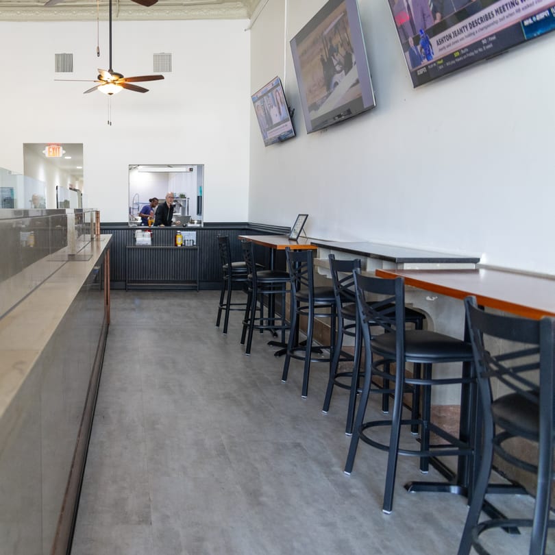 Bright restaurant interior with a service counter, bar stools, an open kitchen, and multiple wall-mounted TVs.