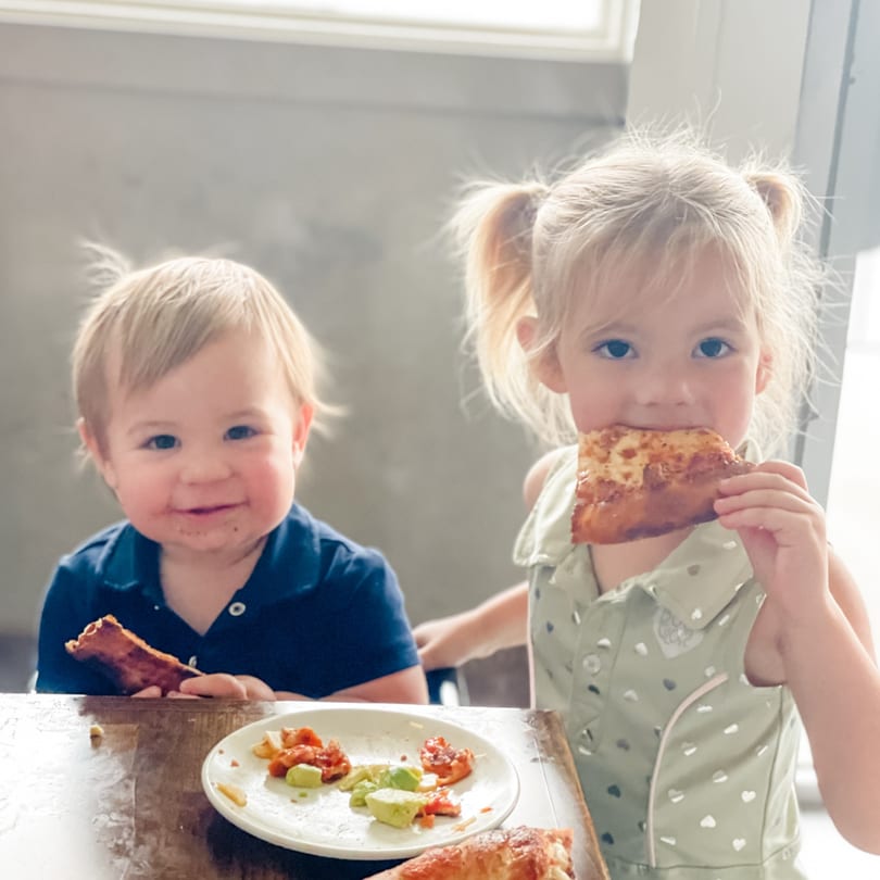 Two young children happily eating pizza and other food at a table, with messy faces.