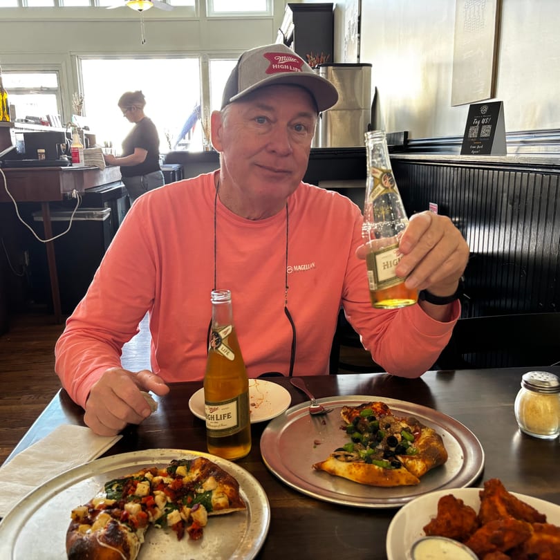 Smiling man with Miller High Life beer, pizza, and wings at a restaurant table.