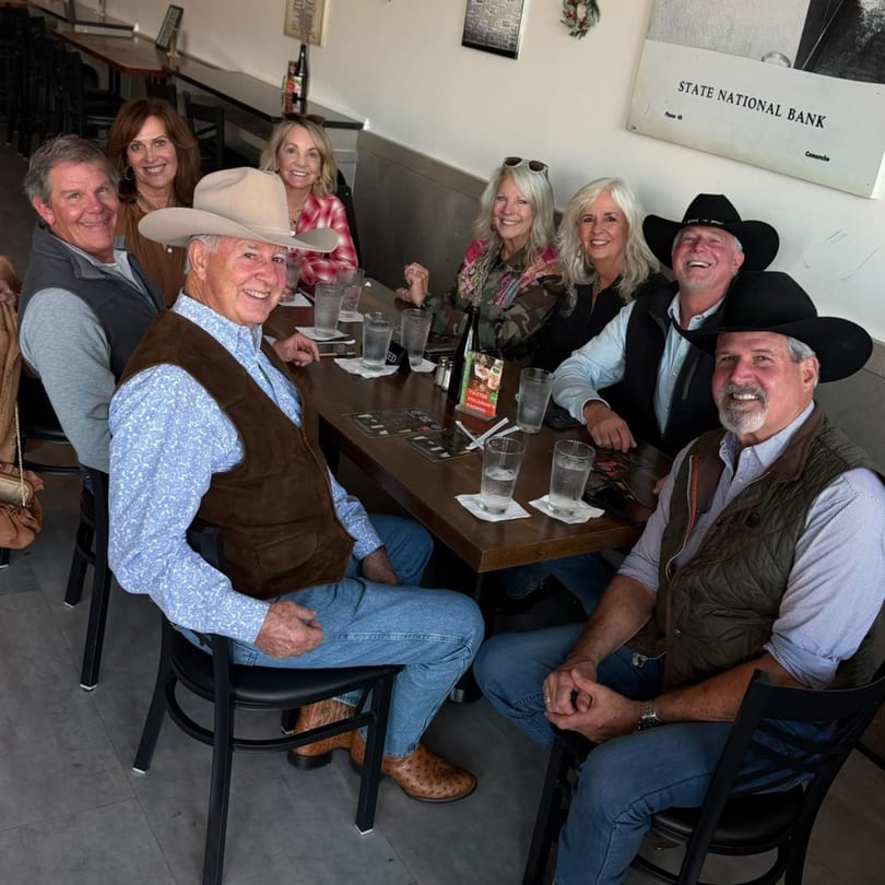 Eight smiling adults, some in cowboy hats, gather around a table in a restaurant.