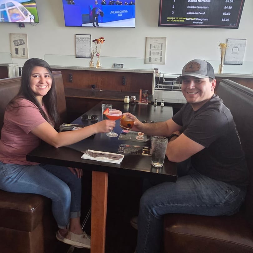 Smiling couple toasting drinks at a restaurant booth with TVs in the background.