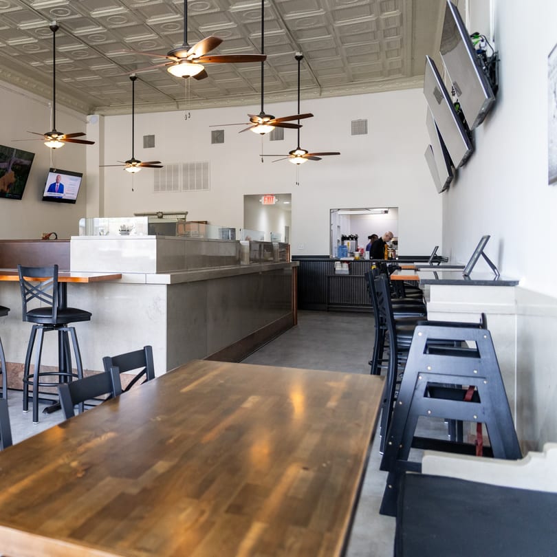 Clean, modern restaurant interior with wooden tables, black chairs, a service counter, and ceiling fans.