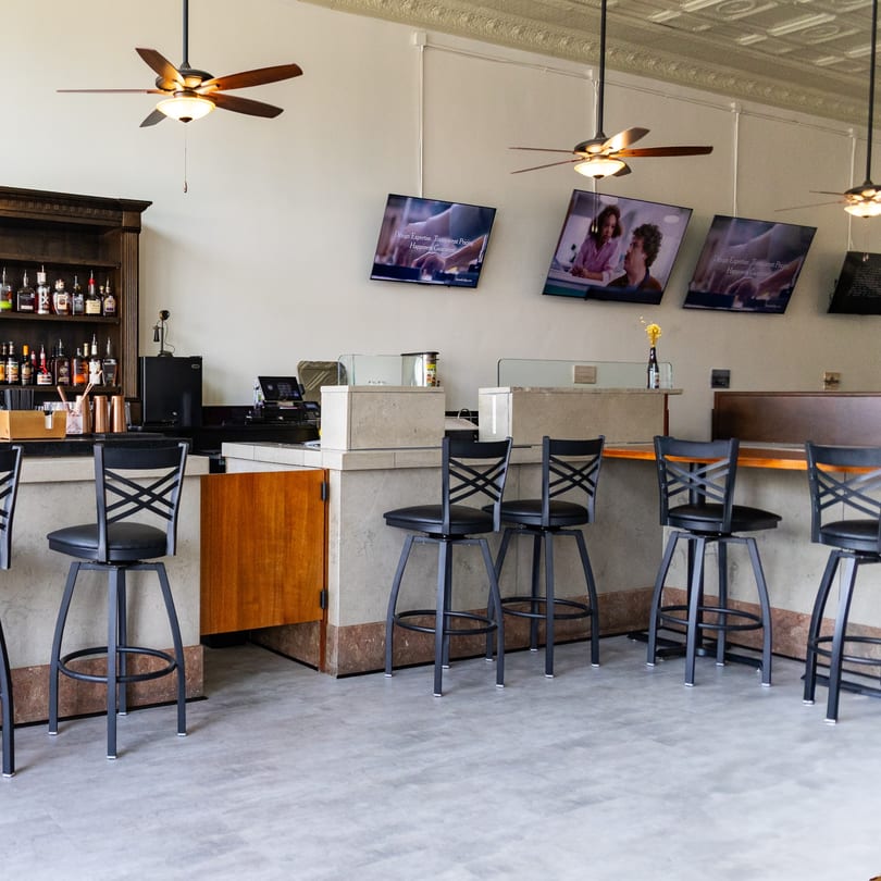 Modern bar area with black stools, liquor bottles, digital screens, and ceiling fans.