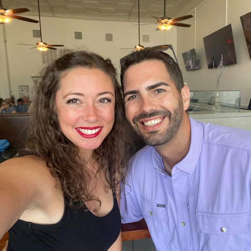 Smiling couple posing in a restaurant, with ceiling fans and TVs visible behind them.