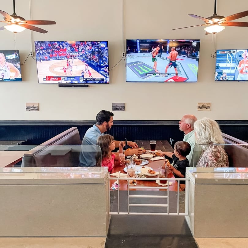 Family eating pizza in a restaurant booth with multiple sports games playing on wall-mounted TVs.