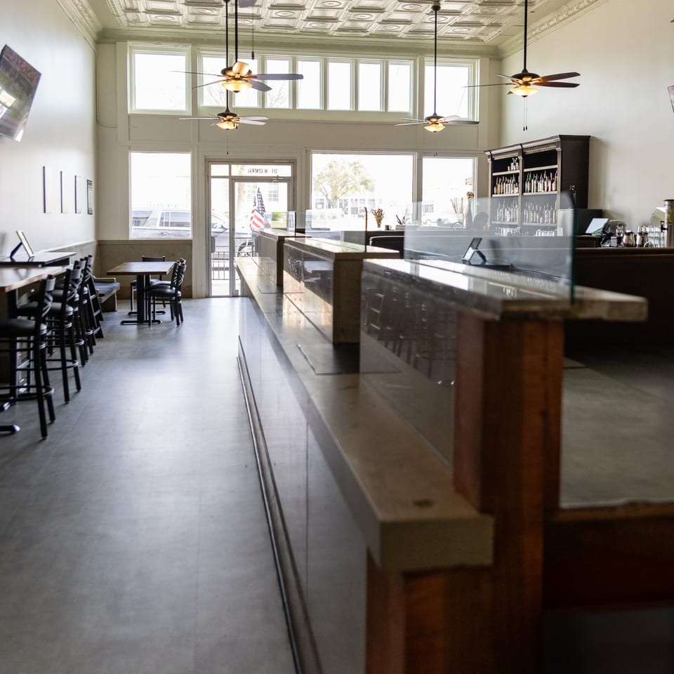 Bright, empty restaurant interior featuring a long bar, tables, chairs, and decorative tin ceiling with fans.