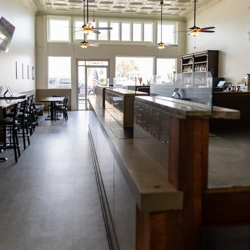 Bright, empty restaurant interior featuring a long bar, tables, chairs, and decorative tin ceiling with fans.