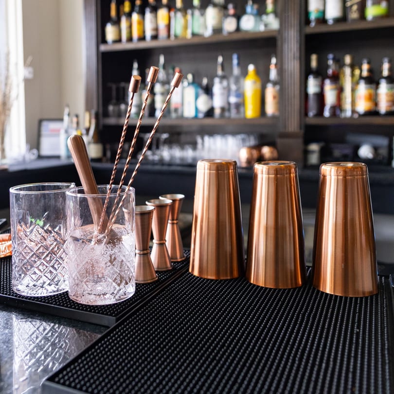 Copper bar tools and cocktail glasses on a black mat, with liquor bottles blurred in the background.