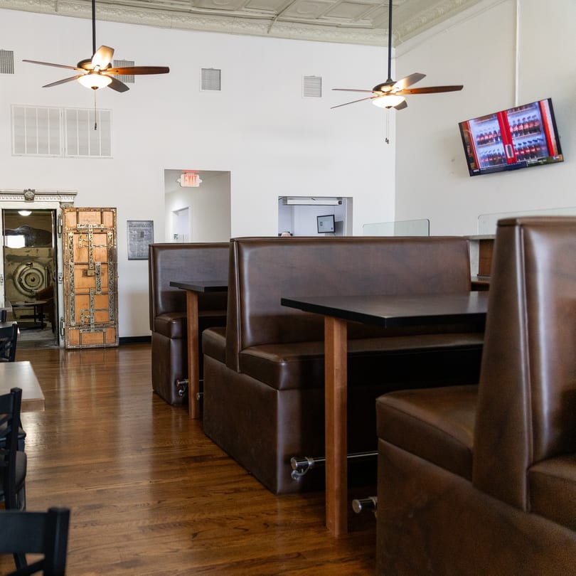 Restaurant dining area with brown leather booths, wooden tables, hardwood floors, and an old bank vault door.