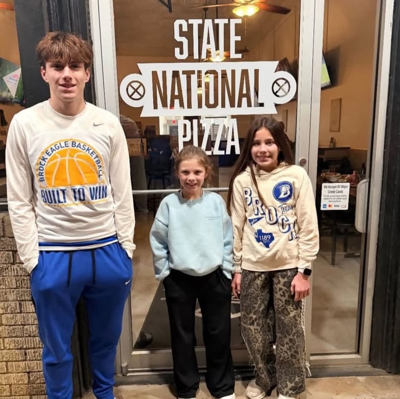 Three smiling people, a young man and two girls, stand outside State National Pizza.
