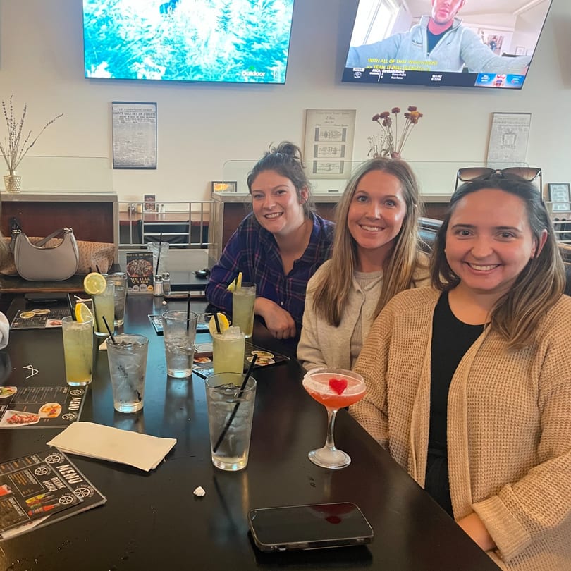 Three smiling women sit at a restaurant table with cocktails and menus.